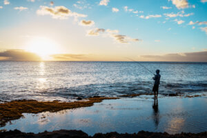 An unrecognizable man stands in the shallow waters of the ocean, holding a fishing pole as he enjoys a sunset on the beaches of the Pacific Rim.