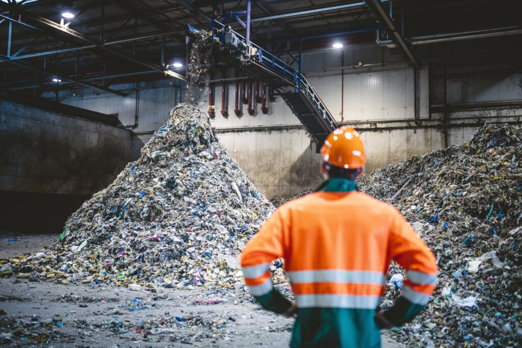 Rear view of young male worker in helmet, pollution mask, and reflective clothing observing waste falling from conveyor belt onto pile at facility.