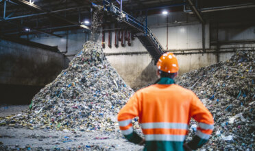 Rear view of young male worker in helmet, pollution mask, and reflective clothing observing waste falling from conveyor belt onto pile at facility.