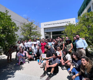 Dozens of smiling activists pose for a photo outside of South Coast Air Quality Management District Headquarters, two activists are holding signs which read “no more deaths” and “clean air now.” They are surrounded by trees and the sky is bright blue.